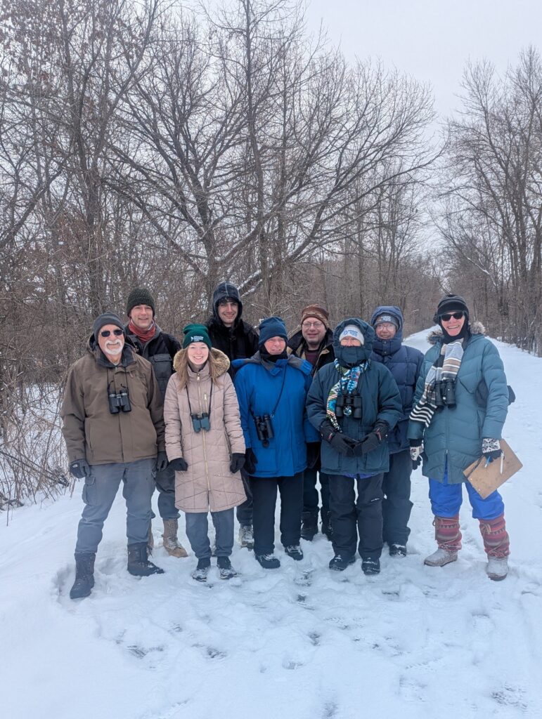 Nine people in winter coats with binoculars on a trail in winter.