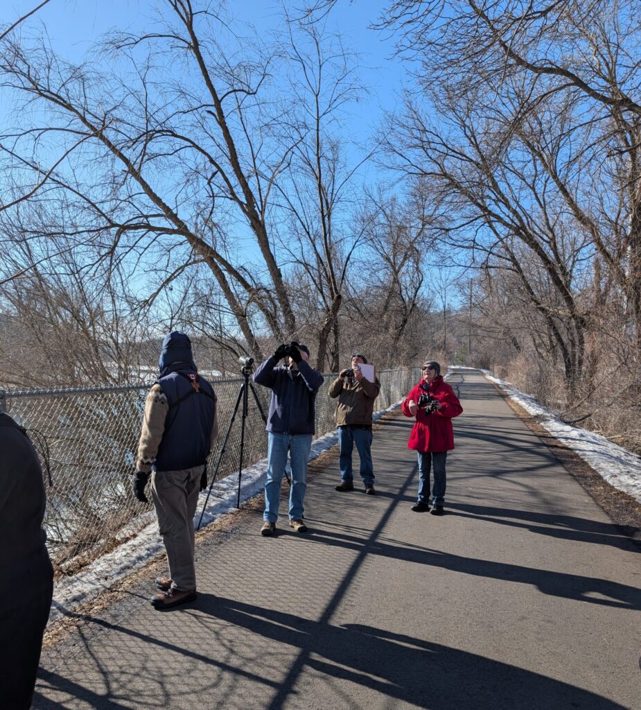 Four people on a paved trail looking up, some through binoculars.